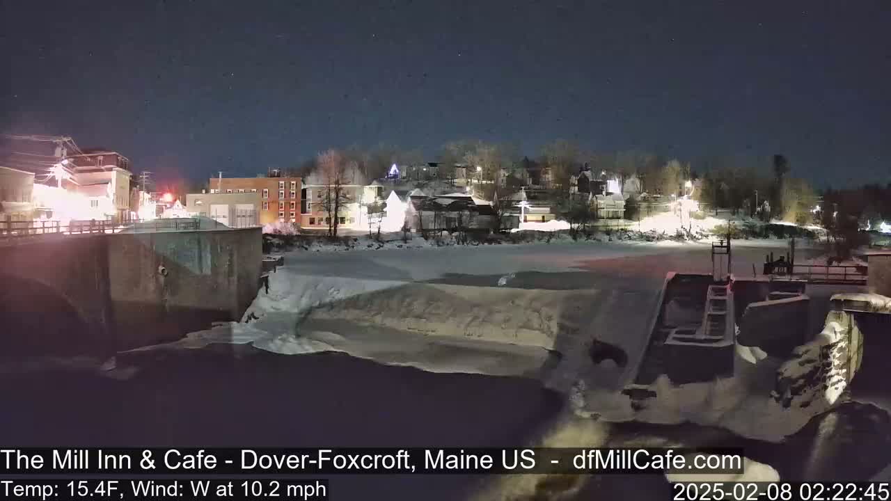 A snow-covered town at night is seen from across a partially frozen river, with a 10.2 mph west wind and a temperature of 15.4°F.