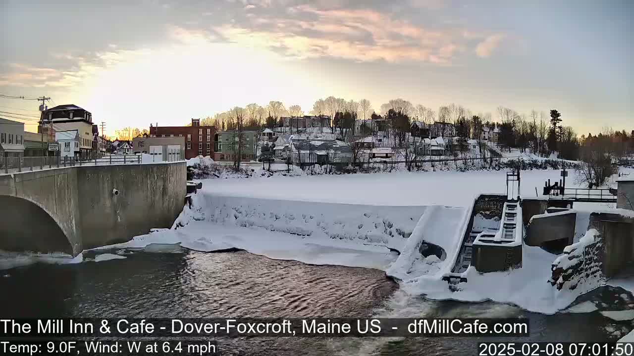 A snow-covered town is seen from a bridge overlooking a partially frozen river and dam at sunrise.