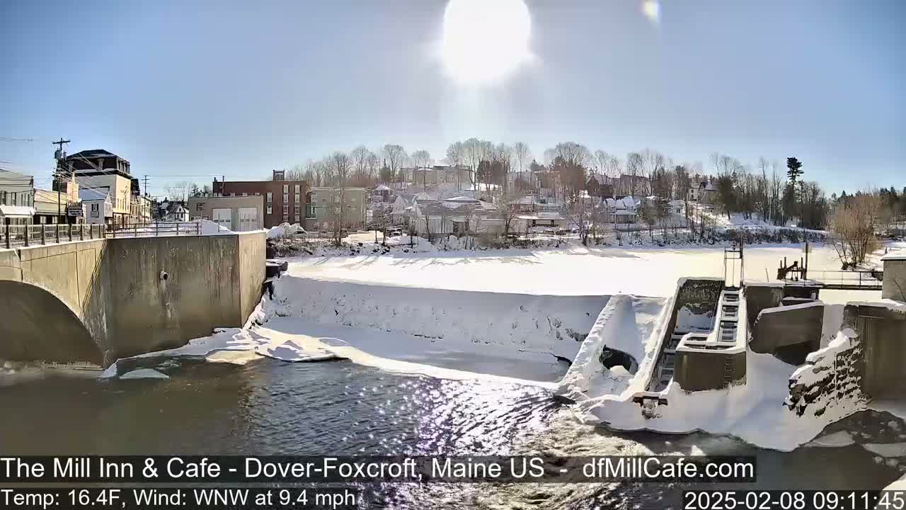 A sunny winter scene shows a snow-covered dam and river, with a small town visible in the background.