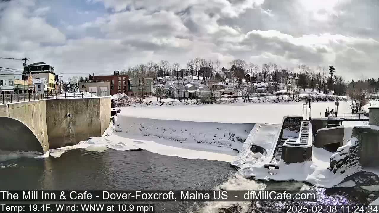 A snow-covered dam and partially frozen river are seen in front of a small town under a cloudy sky.