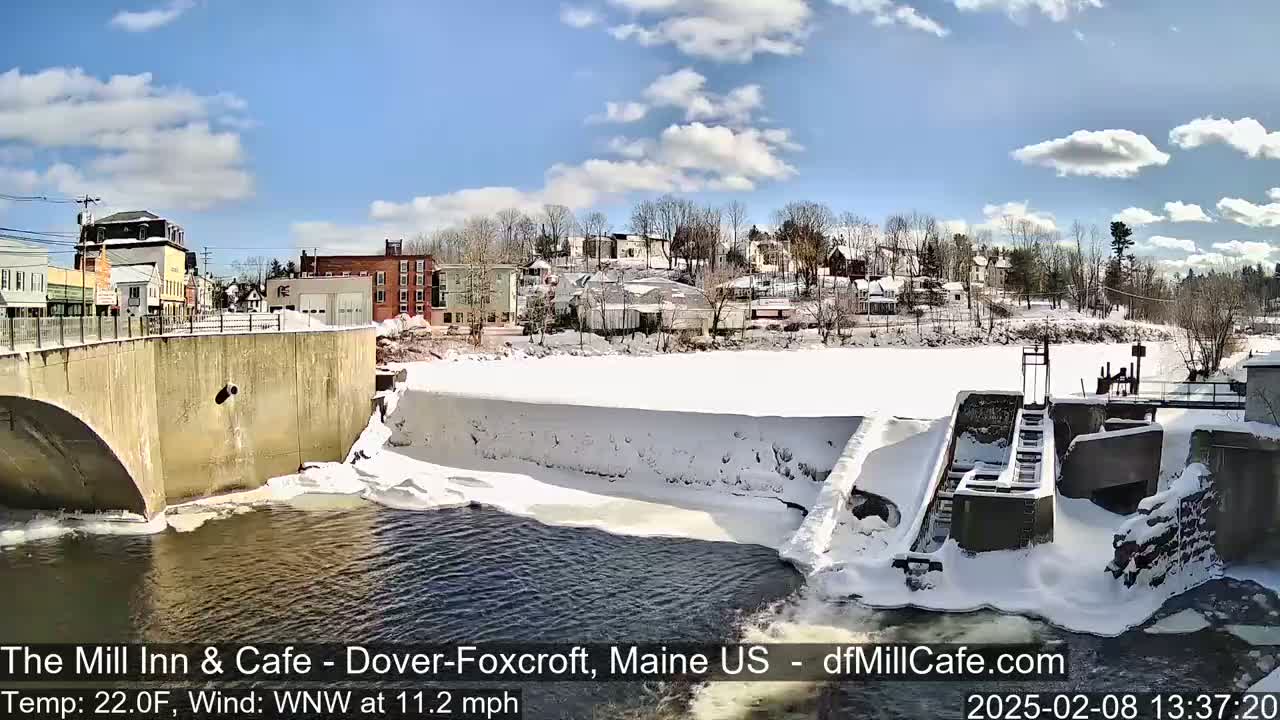 A partially frozen river flows past a snow-covered dam and under a bridge, with a small town visible in the background under a partly sunny sky.