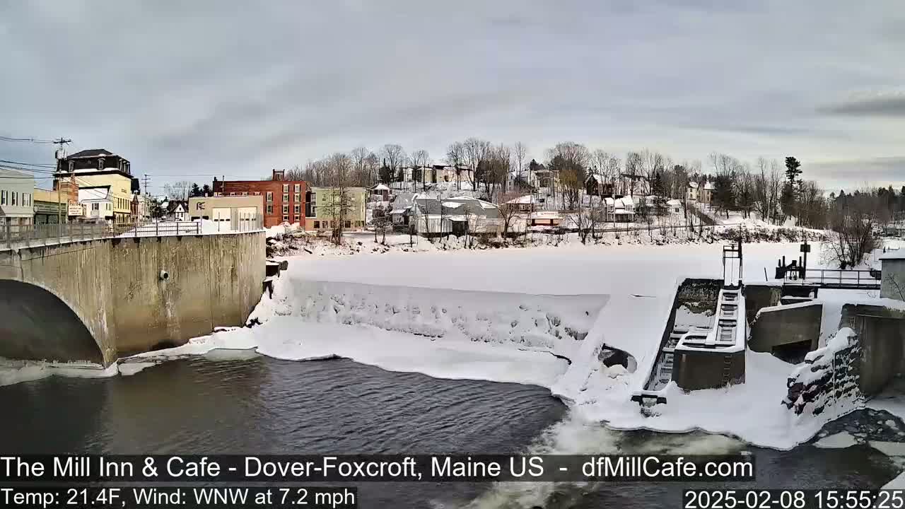 A snow-covered town is seen from a bridge overlooking a partially frozen river with a dam and a low-water flow.