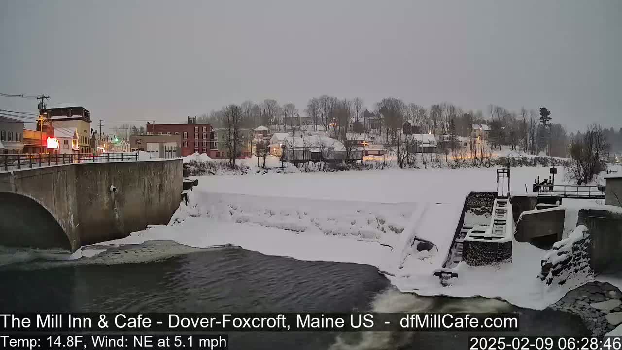 A snow-covered town is viewed from a bridge over a partially frozen river with a dam and a low light overcast sky.
