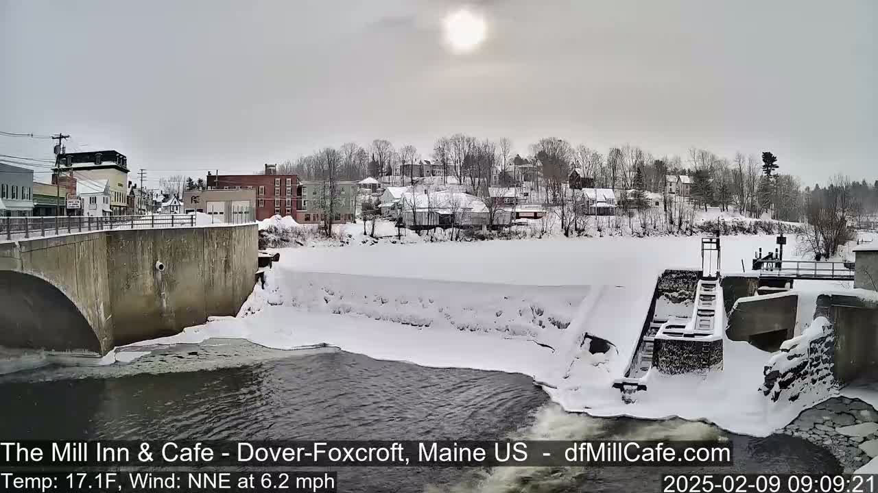 A snow-covered town is seen from a bridge overlooking a partially frozen river and dam, under a bright, overcast sky.