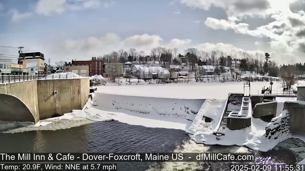 A snow-covered dam and river are seen, with a small town in the background under a partly cloudy sky.