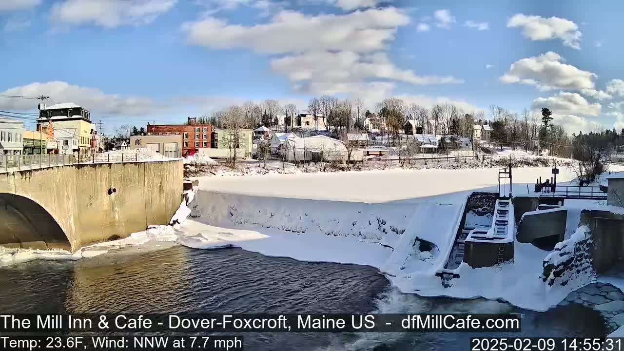 A snow-covered town sits beside a partially frozen river with a dam and bridge under a partly cloudy sky.