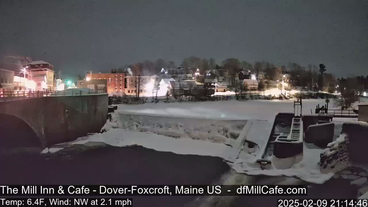 A nighttime view of a snow-covered town and a partially frozen river with a dam, under a dark sky with a light Northwest wind.