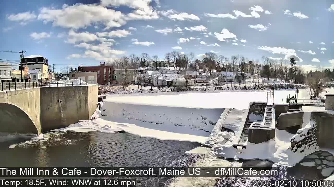 A partially frozen river flows past a snow-covered dam and town under a partly cloudy, sunny sky.