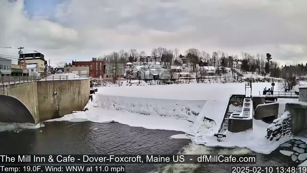 A snow-covered town with a river partially frozen over, featuring a dam and bridge, under a cloudy sky.