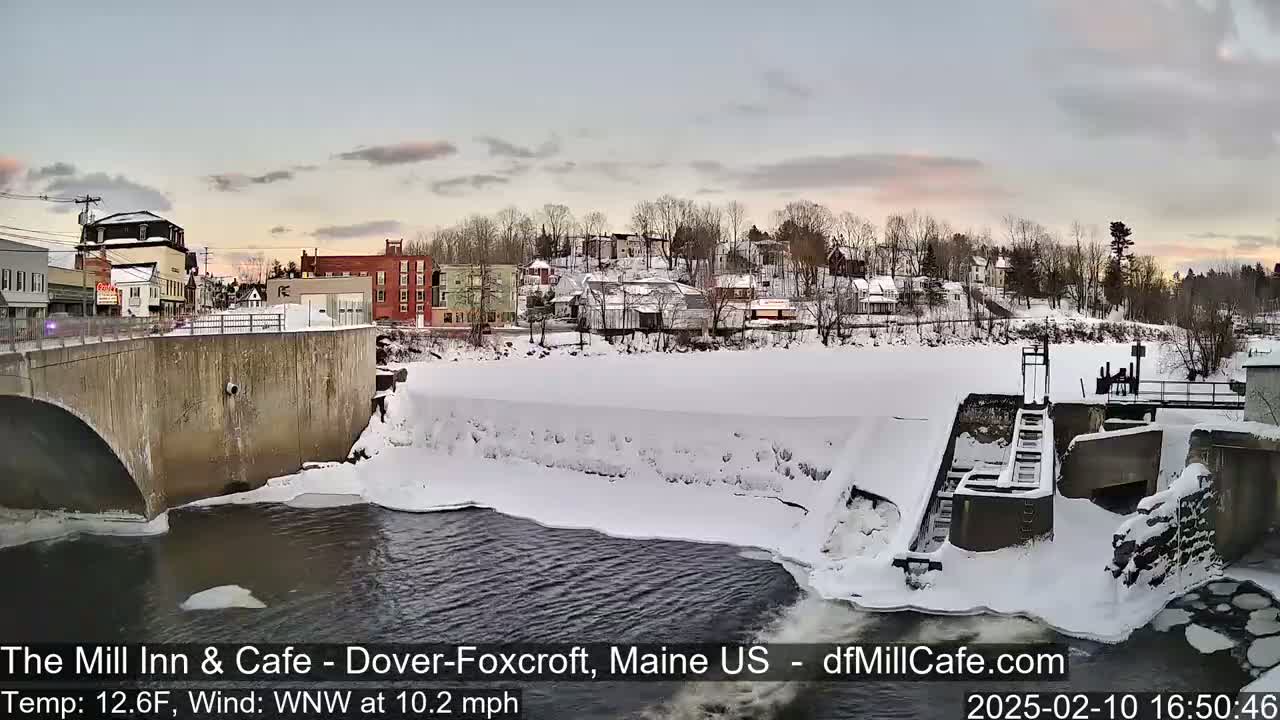 A snow-covered town sits beside a partially frozen river flowing over a dam, under a twilight sky.