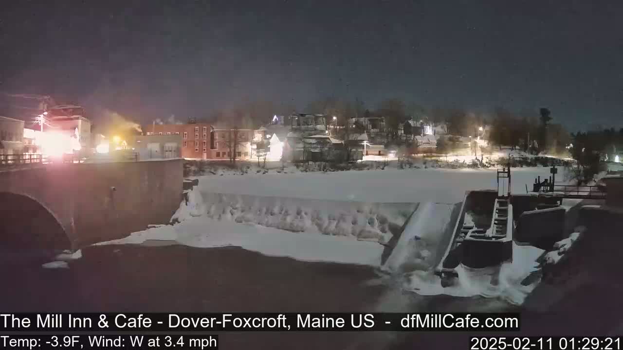 A nighttime view of a snow-covered river flowing past a dam and town buildings under a dark sky with a slight west wind at 3.4 mph and a temperature of -3.9°F.
