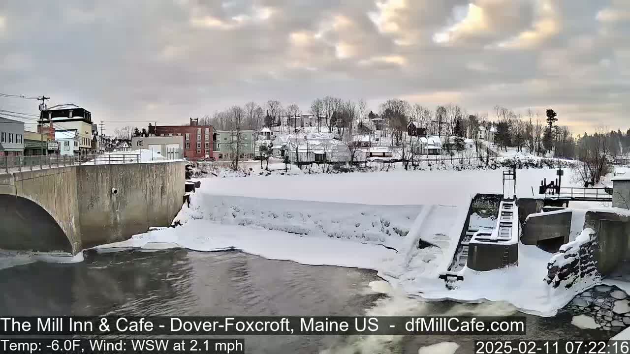 A snow-covered town sits beside a partially frozen river with a dam and lock system, under a cloudy sky with hints of sunrise.