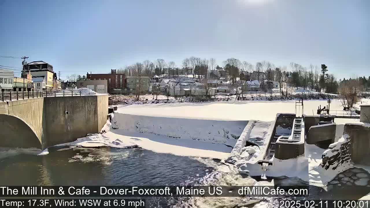A snow-covered dam and river are seen, with a town in the background under a sunny sky.