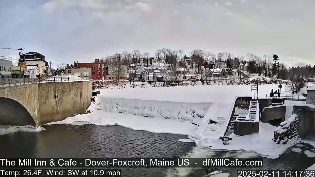 A snow-covered dam and river flow past a small town under a cloudy sky.
