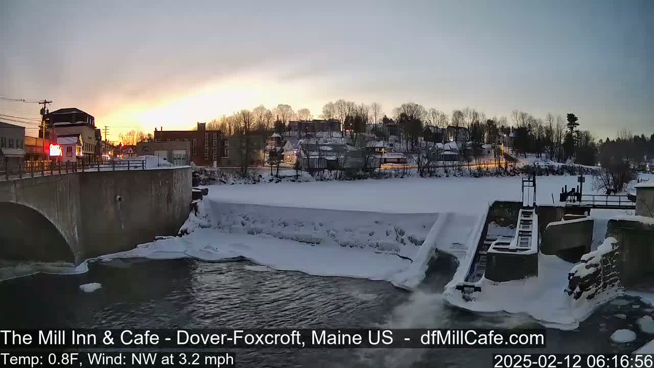 A snow-covered river flows past a dam and under a bridge, with a small town visible in the background under a twilight sky.