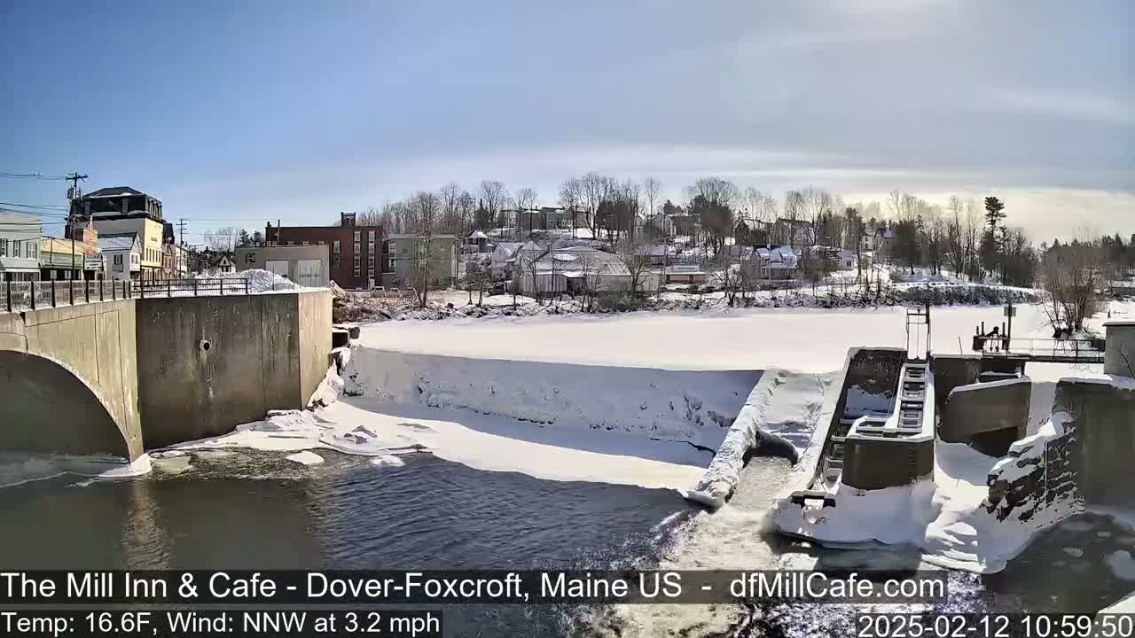 A partially frozen river flows past a snow-covered dam and town under a sunny sky.