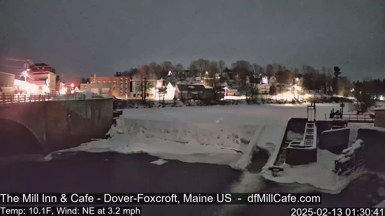 A nighttime view of a snow-covered river and town, with a partially frozen river flowing past a dam under a dark sky.