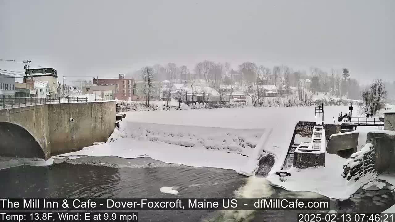 A snow-covered town and river with a dam and bridge are seen during a snowfall with an easterly wind.