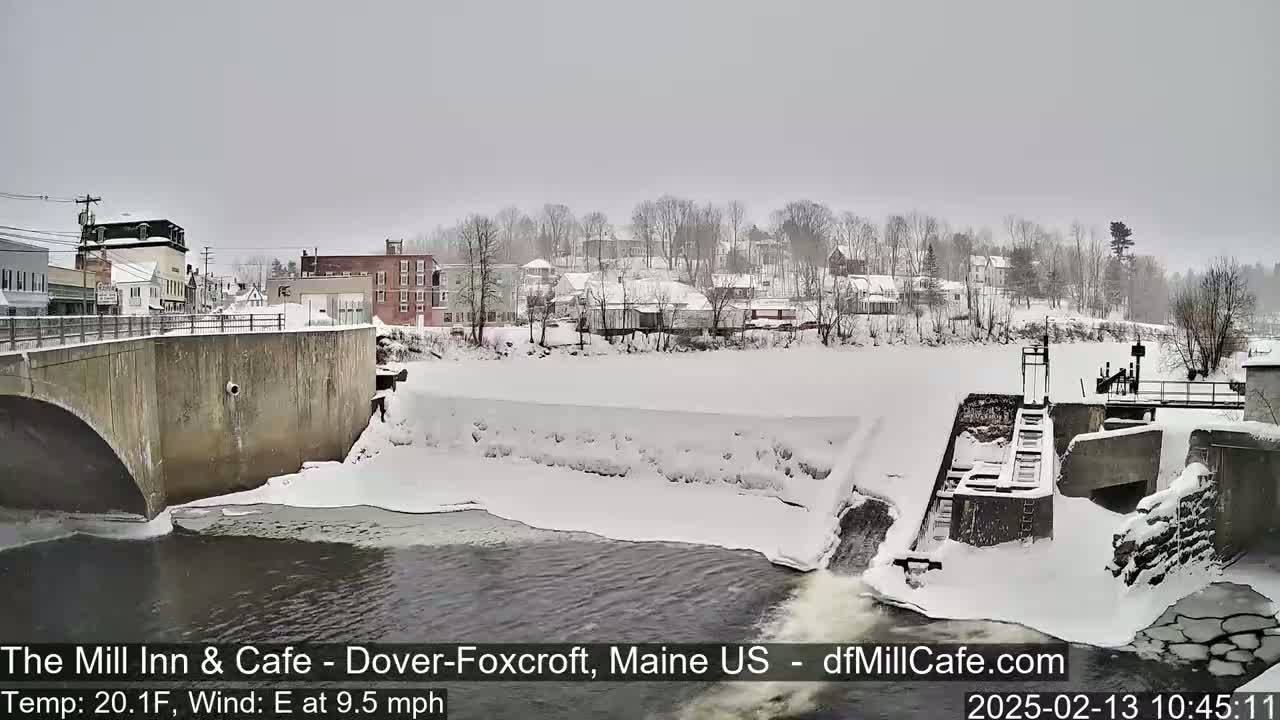 A snow-covered town is seen from a bridge overlooking a partially frozen river and a dam, with a light snowfall.