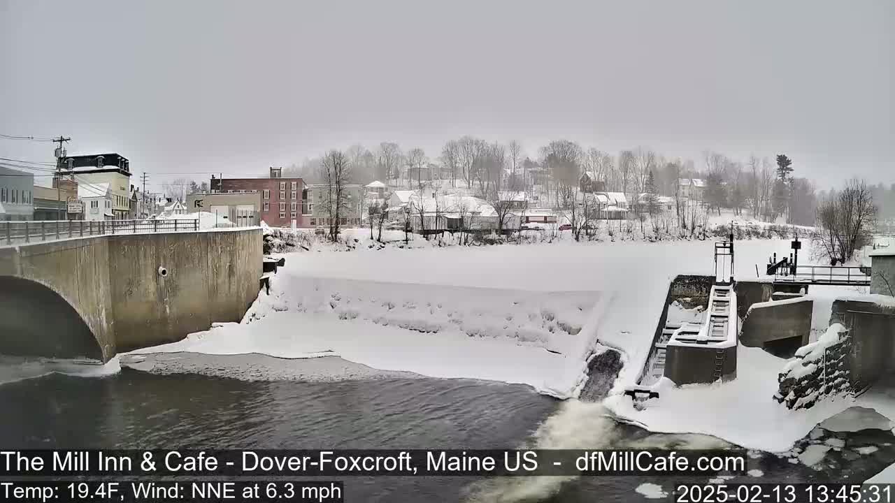 A snow-covered town and river scene features a bridge, a dam, and buildings under an overcast sky.