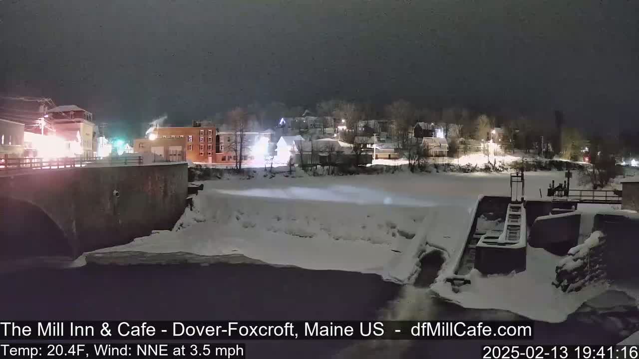 A nighttime view of a snow-covered town by a partially frozen river, with a dam and bridge visible under a dark sky.