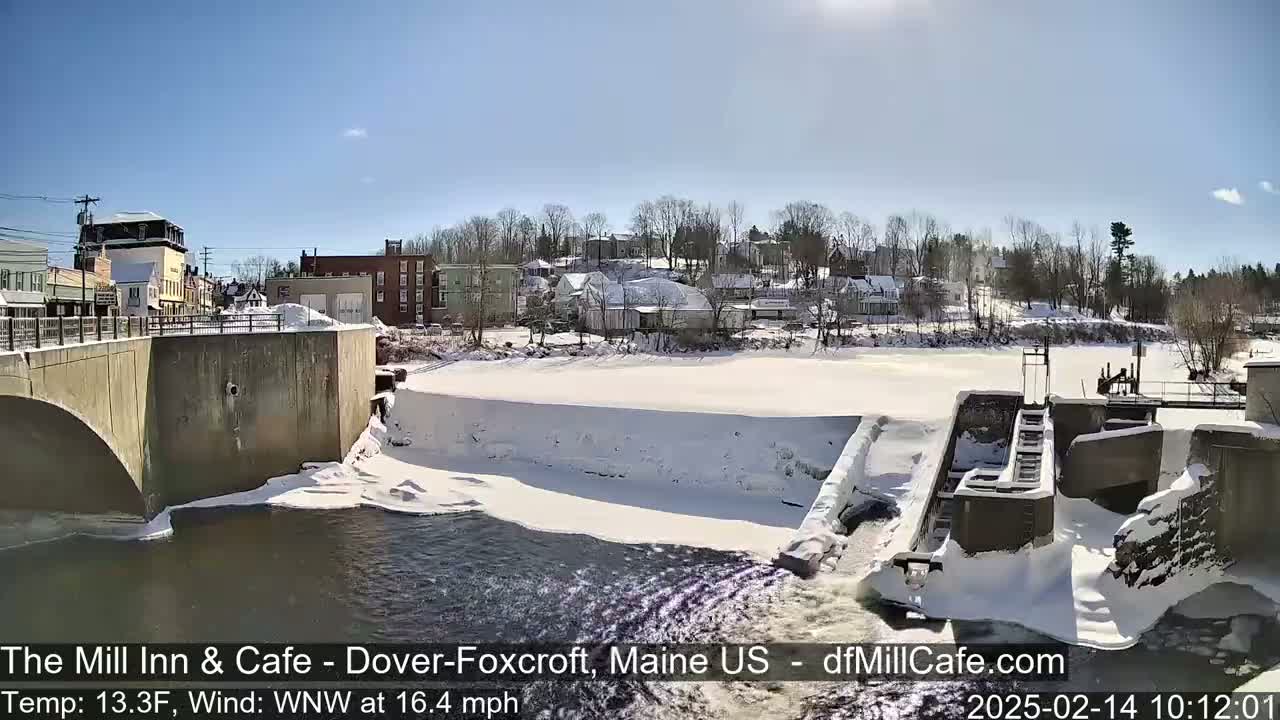 A snowy river flows past a dam and a town under a clear, sunny sky.