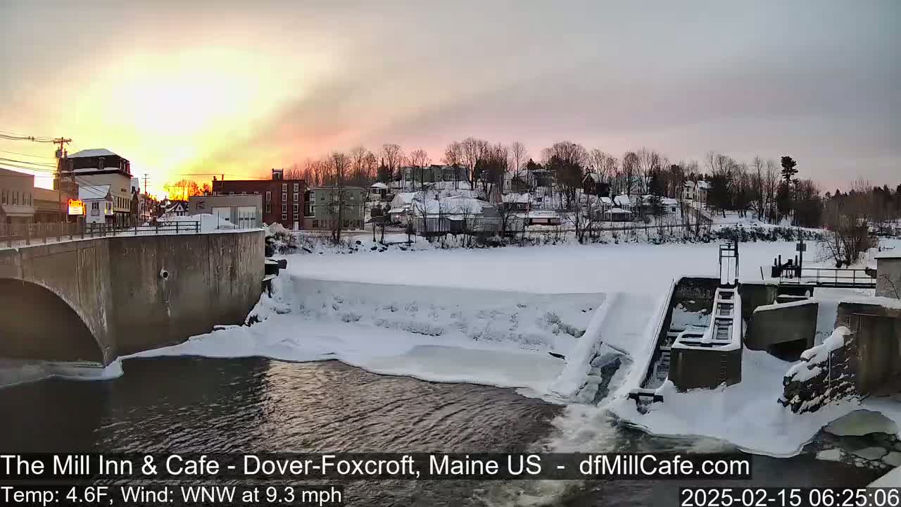 A snow-covered town at sunset is viewed from a bridge overlooking a partially frozen river with a dam.