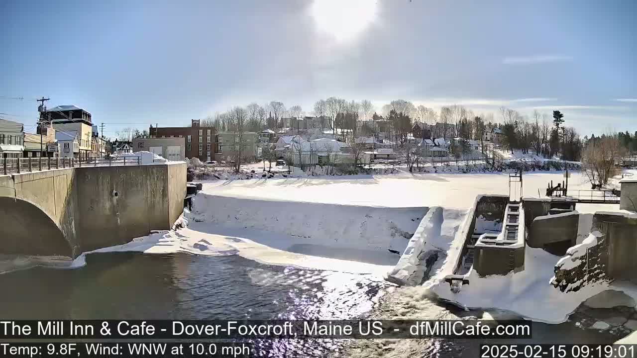 A snowy, sunny day reveals a town's buildings and a dam partially covered in ice and snow, with a river flowing beneath a bridge.