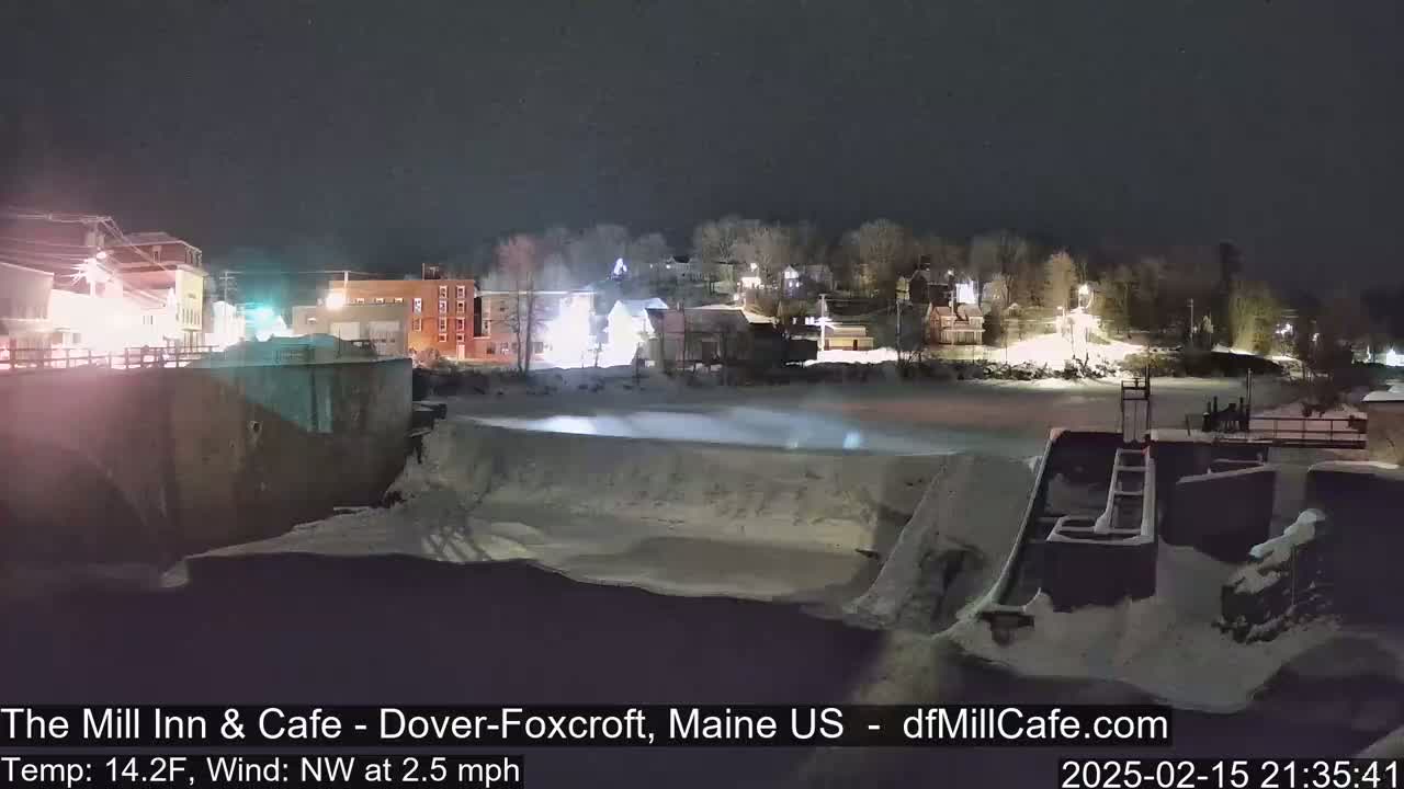 A nighttime view of a snow-covered town near a river, with a cold temperature and a light northwest wind.