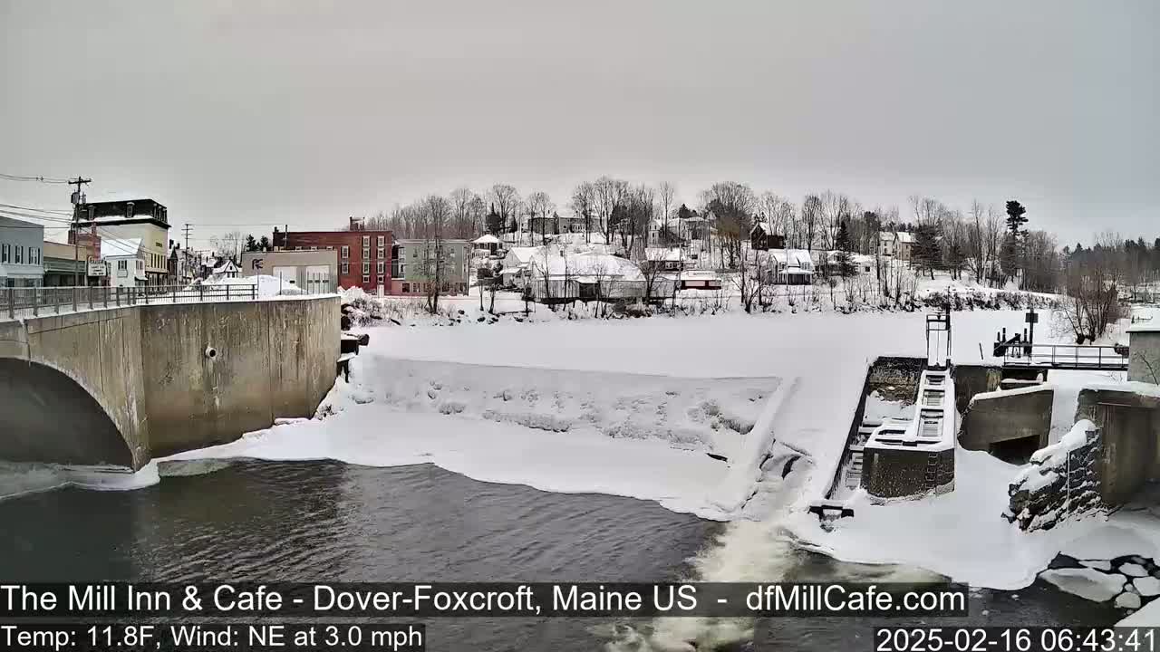 A snow-covered town is seen from across a river with a dam and partially frozen water under an overcast sky.