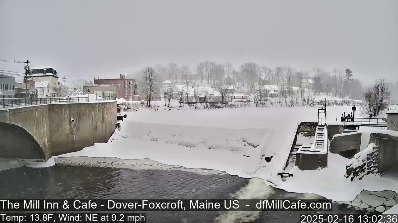 A snow-covered town is seen from a bridge overlooking a dam and a partially frozen river during a light snowfall with a northeast wind.