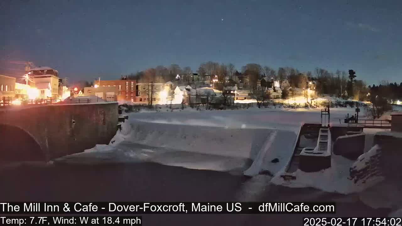 A snow-covered town at night, with a partially frozen river and dam visible in the foreground, under a clear, dark sky with a west wind at 18.4 mph and a temperature of 7.7°F.