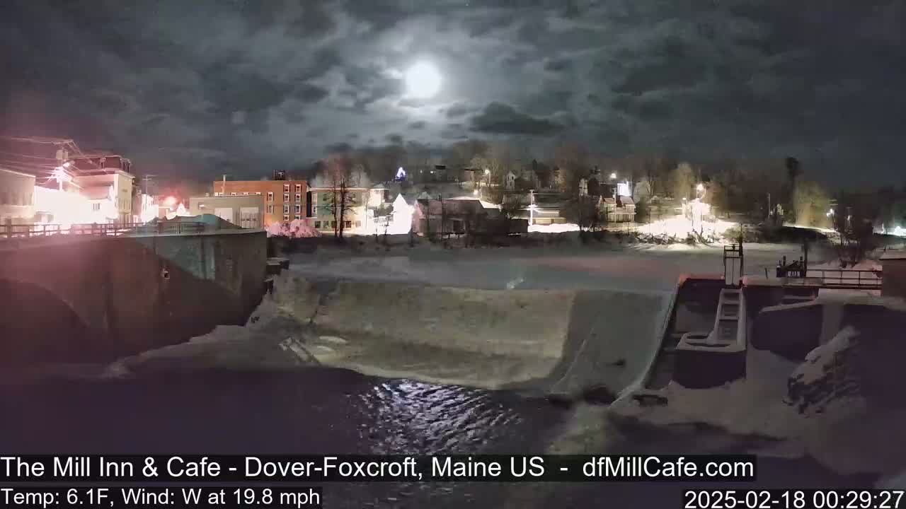 A snow-covered town is seen at night under a partly cloudy sky with a bright moon, next to a river with a dam and a bridge.