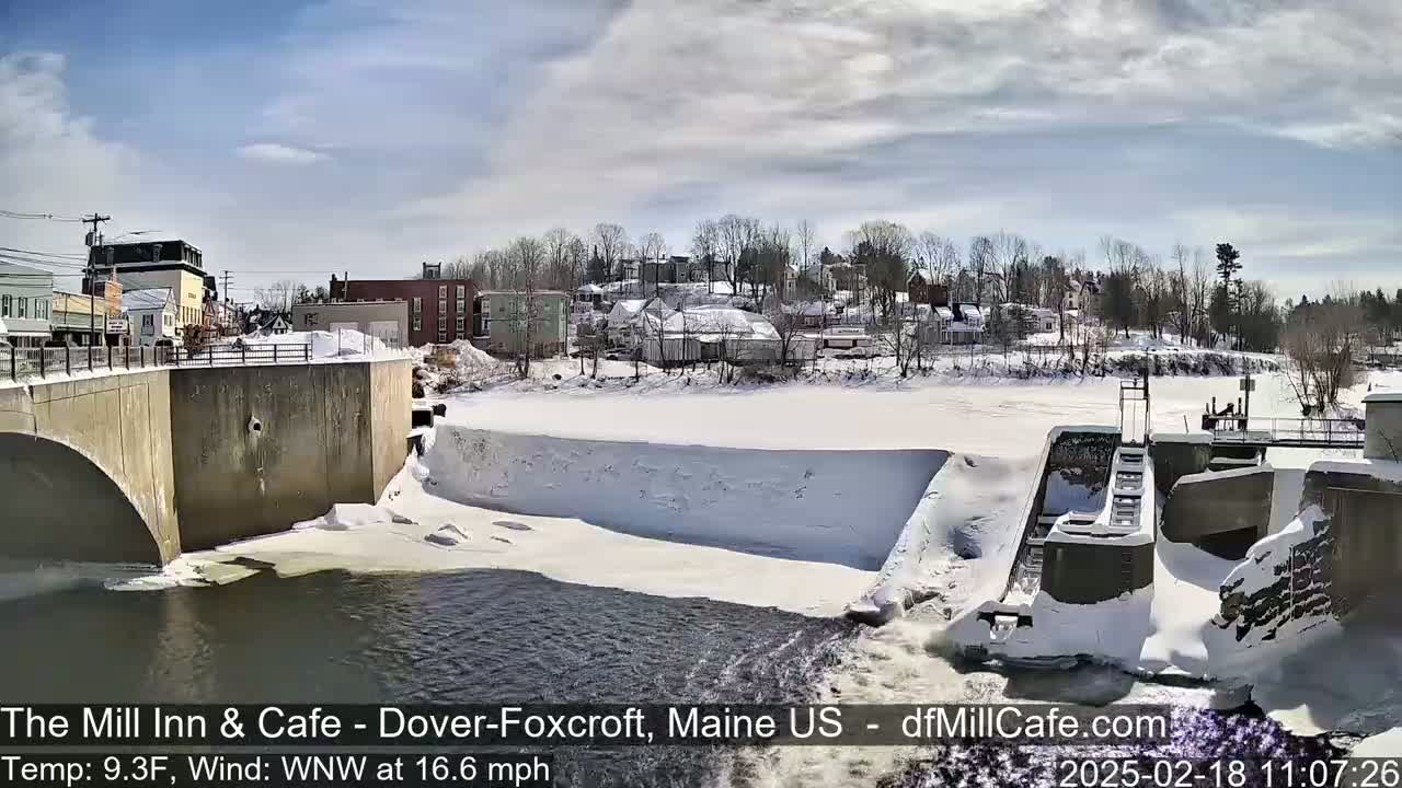A snow-covered dam and river are seen from a bridge, with a small town in the background under a mostly sunny sky.