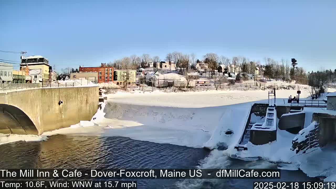 A snow-covered town is seen from across a partially frozen river, with a dam and bridge visible in the foreground under a sunny sky.