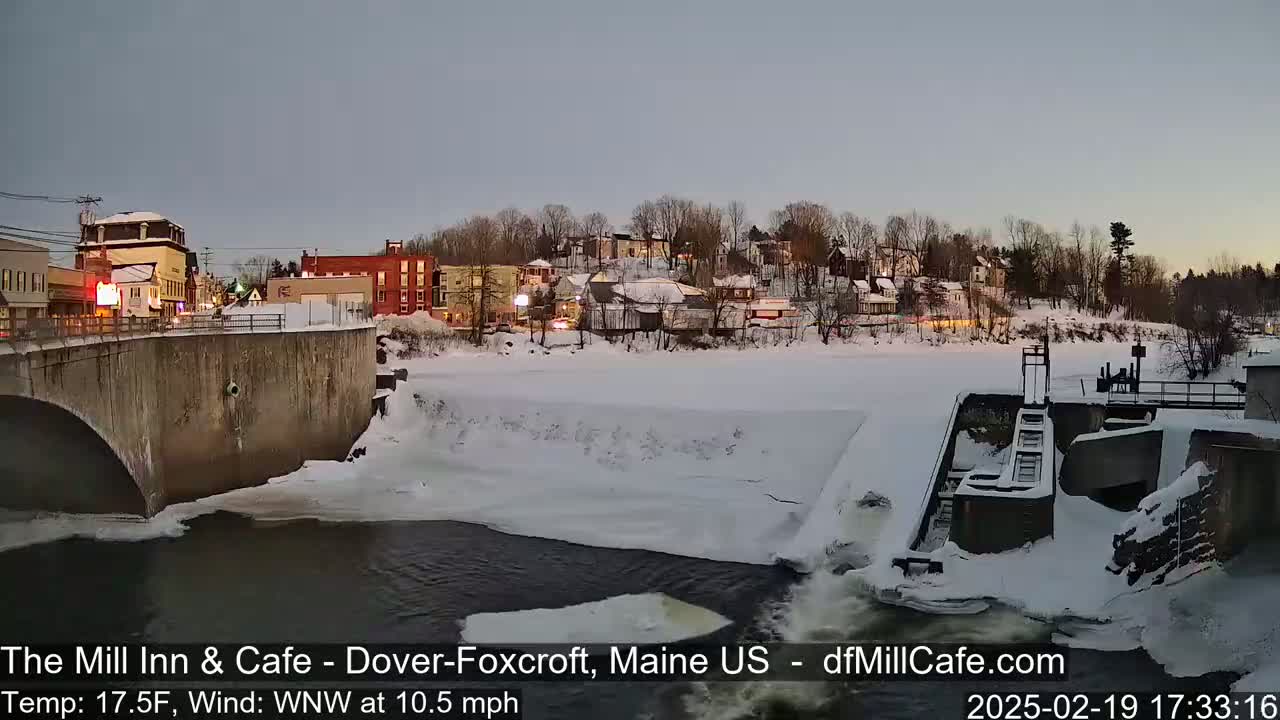 A snow-covered town is seen from a bridge overlooking a partially frozen river with a dam, under a twilight sky.