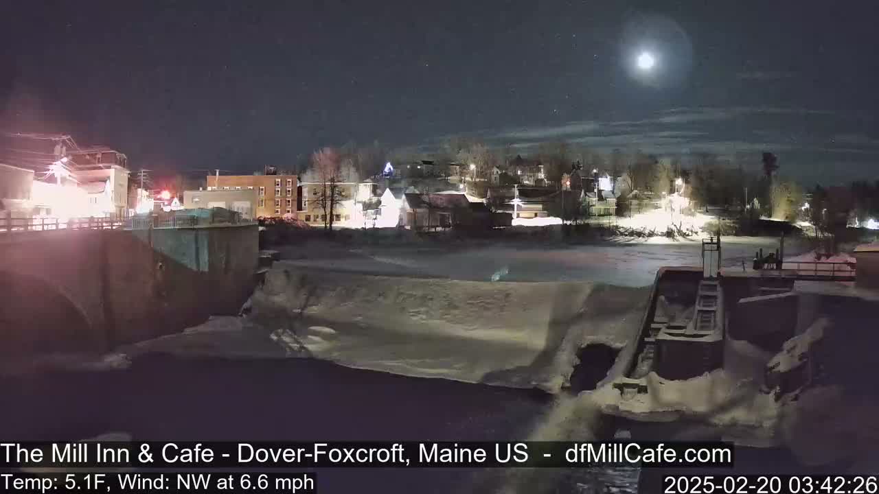 A nighttime view of a snow-covered town with a partially frozen river and a dam under a bright moon.