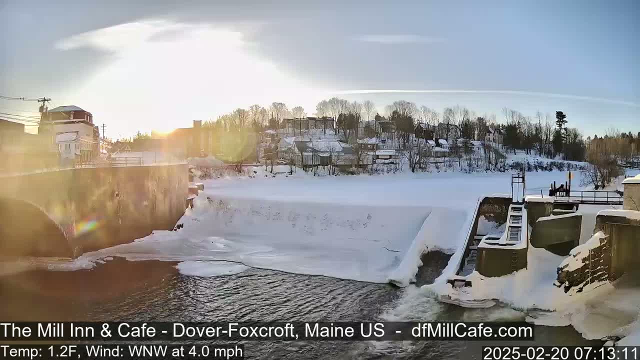 A snow-covered town and river are visible under a sunny sky, with a dam and partially frozen river in the foreground.