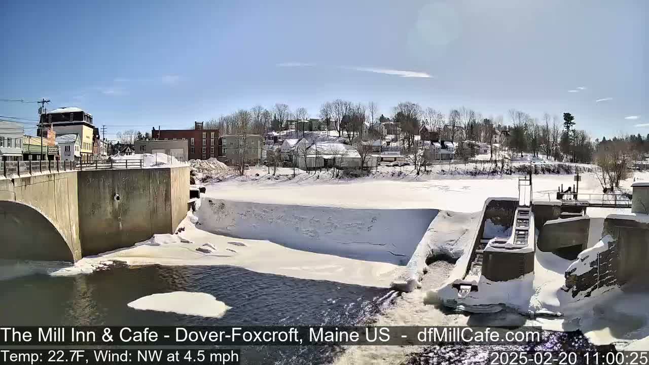 A partially frozen river flows under a bridge near a snow-covered town on a sunny day with a northwest wind.