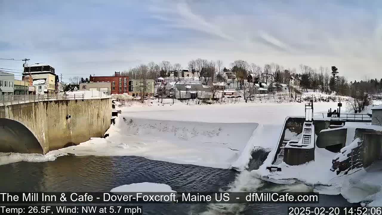A snow-covered town is seen beyond a partially frozen river flowing past a dam on a partly cloudy day.