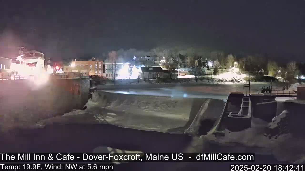 A nighttime view of a snow-covered river flowing past a dam and illuminated buildings, with a northwest wind at 5.6 mph and a temperature of 19.9°F.