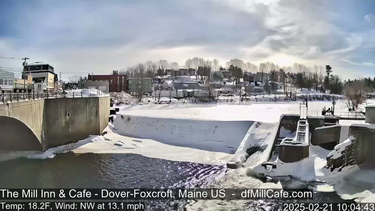 A snow-covered river flows past a dam and a small town under a partly sunny sky.
