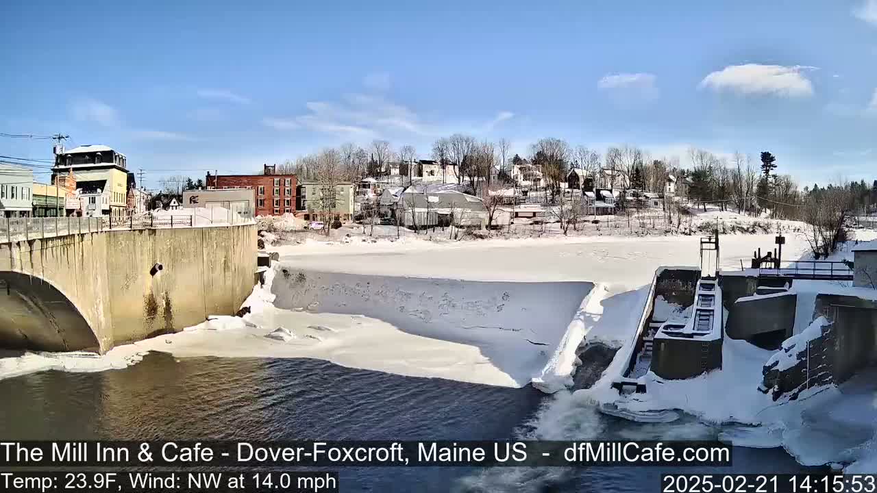 A partly sunny, cold day shows a snow-covered river flowing past a dam and a town with buildings along its banks, a bridge is visible in the foreground.