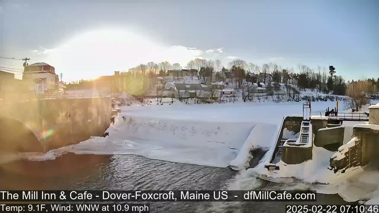 A snow-covered dam and river are seen at sunrise, with a town in the background under a mostly clear sky.