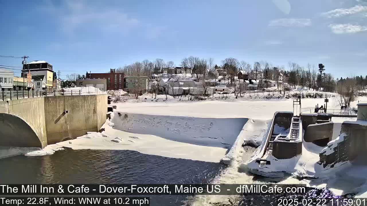 A partially frozen river flows under a bridge past a snow-covered dam and a small town on a sunny, cold day.