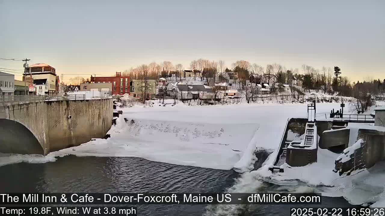 A snow-covered town is seen from above a partially frozen river with a dam and bridge, under a twilight sky.