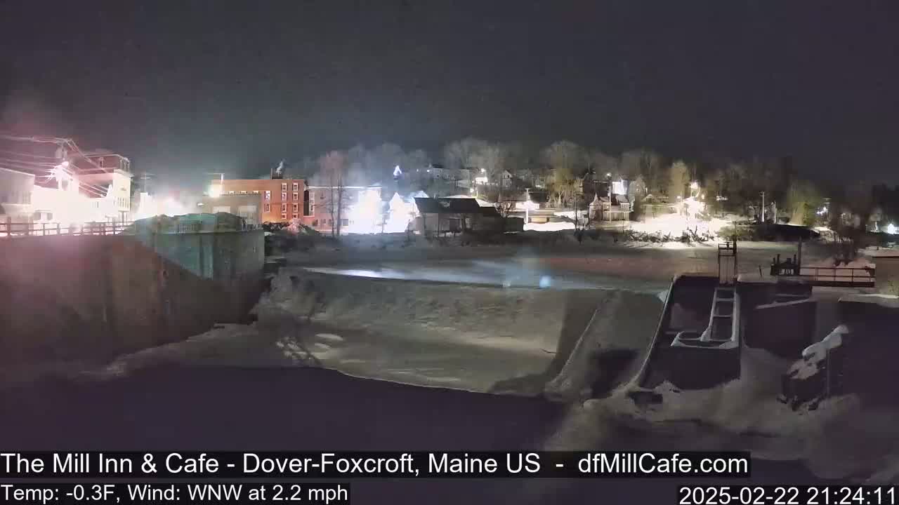 A nighttime view of a snow-covered dam and town, with a cold temperature and light wind.
