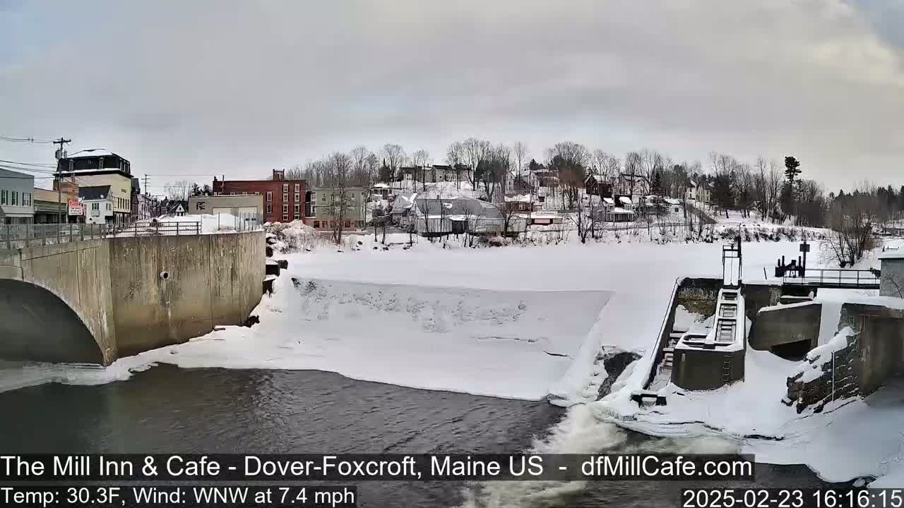 A snow-covered dam and river flow past a small town under a cloudy sky.