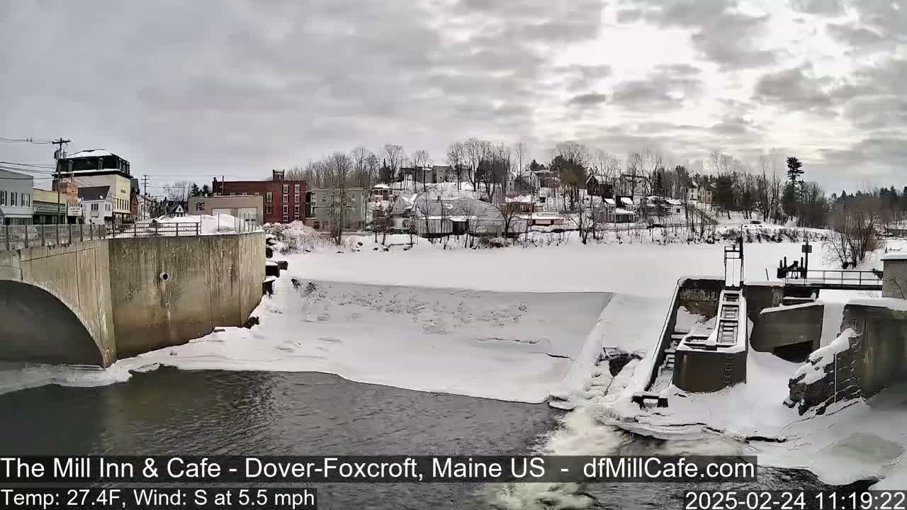 A snow-covered river flows past a dam and under a bridge, with a small town visible in the background under an overcast sky.