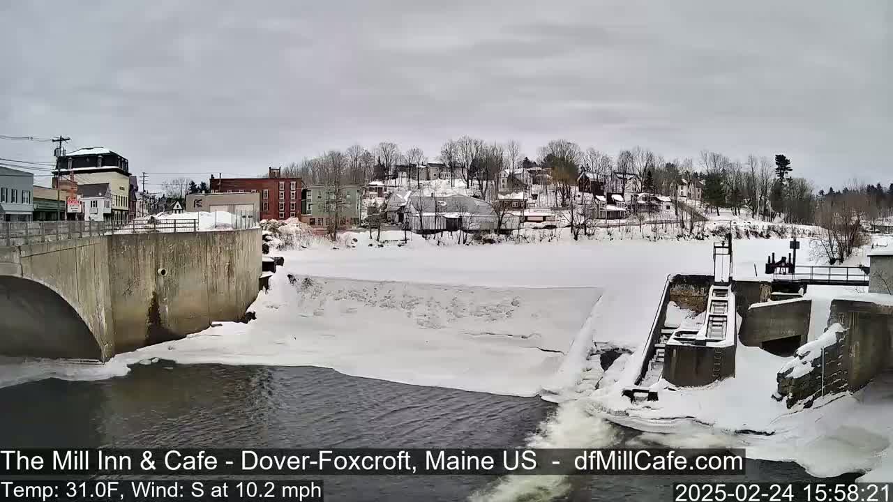 A snow-covered dam and river flow past a small town under an overcast sky.